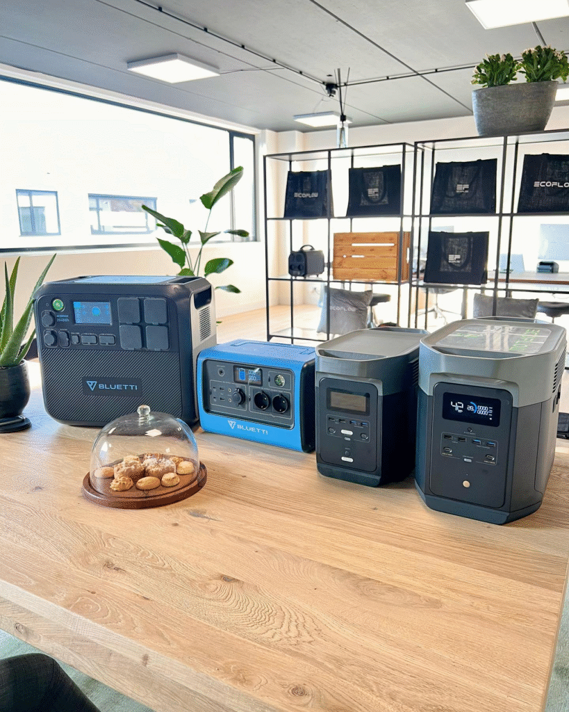 Four portable power stations on a wooden table in an office space with large windows and plants, including black and blue Bluetti models with digital displays and switches