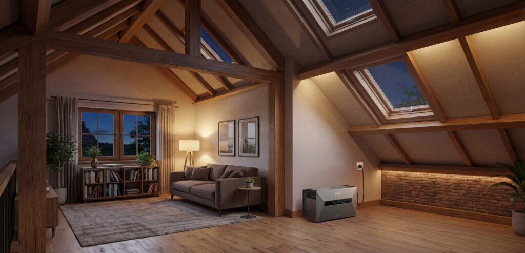 Anker portable power station, used as a small home battery, with a gray casing in a cozy attic room featuring wooden beams, skylights, and warm lighting next to a brick wall, plugged into an outlet for off-grid power.