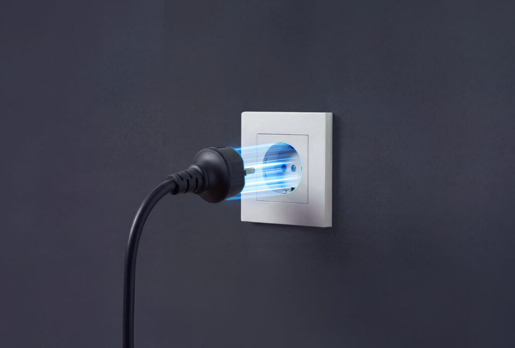 A plug-and-play home battery: a black plug with blue electrical lines plugged into a white outlet on a dark wall, symbolizing energy and electricity.