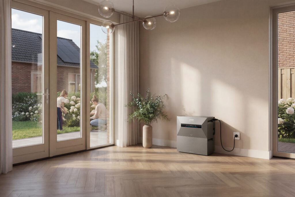 Anker Solix home battery in gray in a modern living room with light wood flooring, with solar panels visible on the roof outside and people in the garden in the background